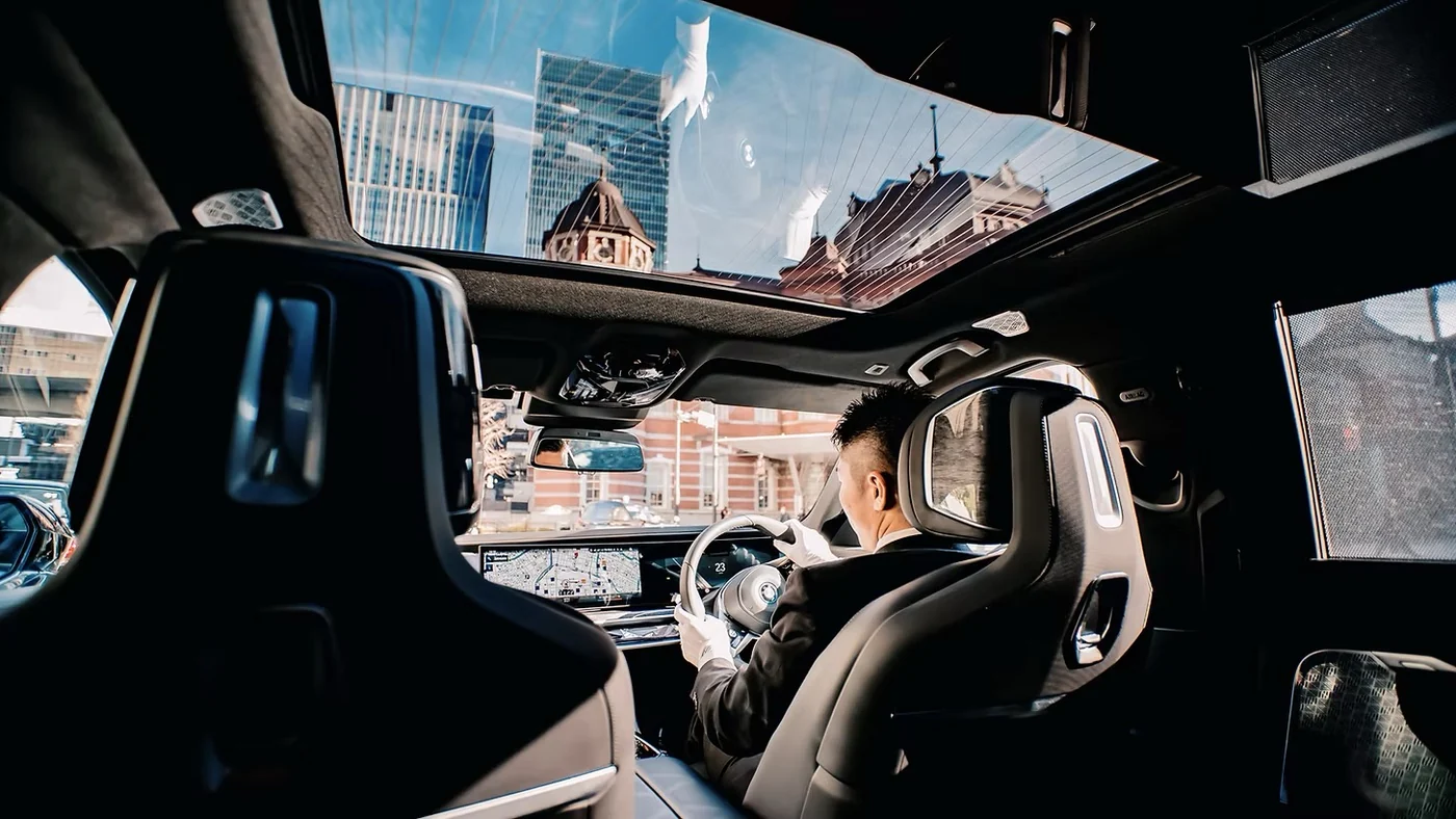 Pandangan dari kursi belakang ke pengemudi bersetelan jas yang mengemudikan BMW, dengan gedung kota terlihat melalui atap panoramik.