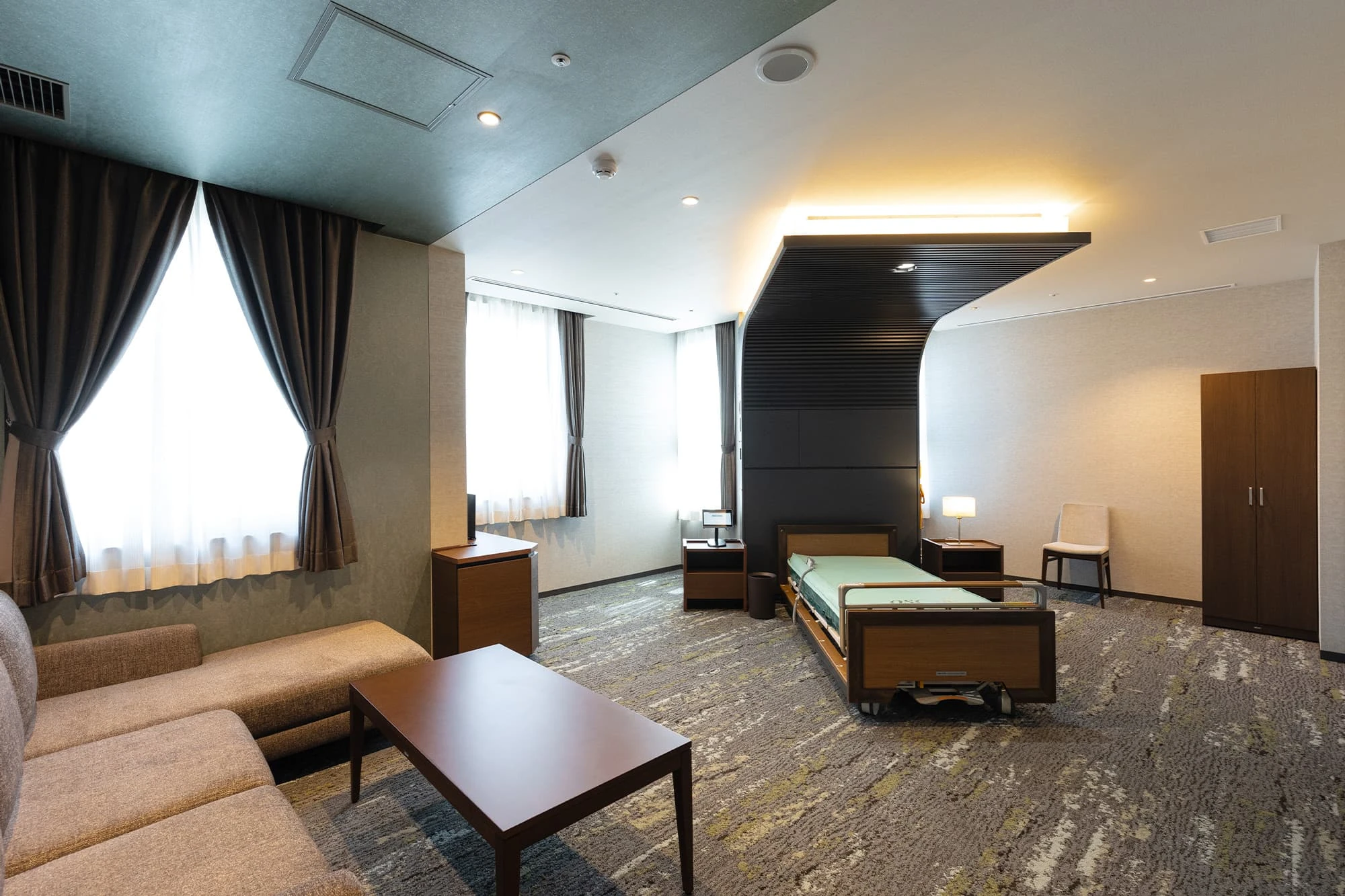 Suite rumah sakit dengan tempat tidur, sofa, meja kopi, jendela besar bertirai, dan lemari kayu.