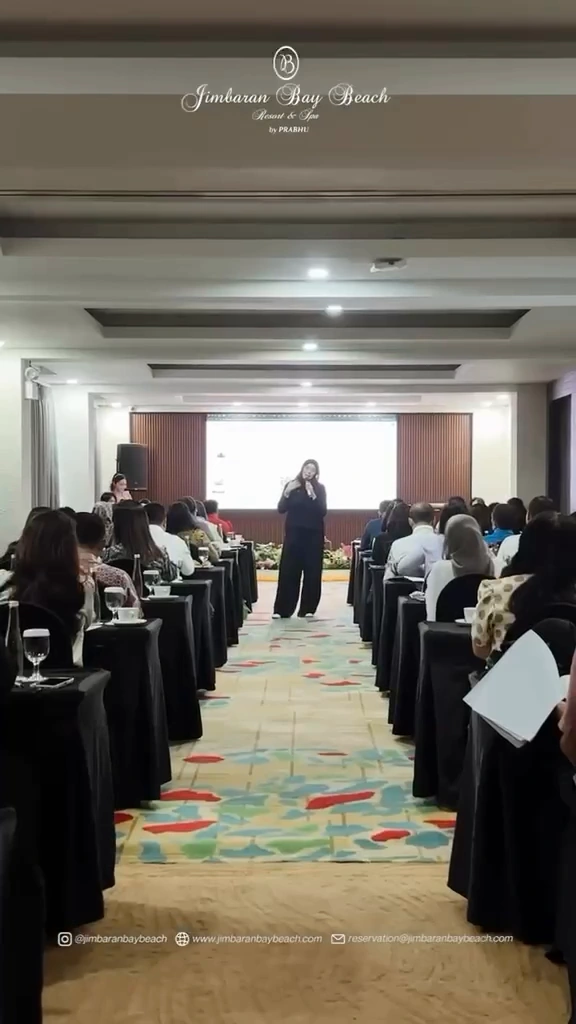 Seorang wanita berbicara menggunakan mikrofon di bagian depan ruang konferensi, dengan peserta seminar duduk di kedua sisi lorong tengah.